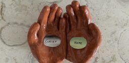 clay sculpture of open hands palms up. One holds card "GRIEF" the other "HOPE"